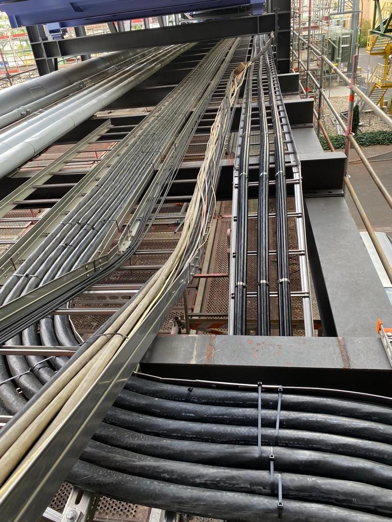 Mehrere Kabeltrassen mit dicken und dünnen Kabeln verlaufen parallel auf einer Metallkonstruktion in einem industriellen Umfeld, umgeben von Gerüsten und technischen Anlagen.