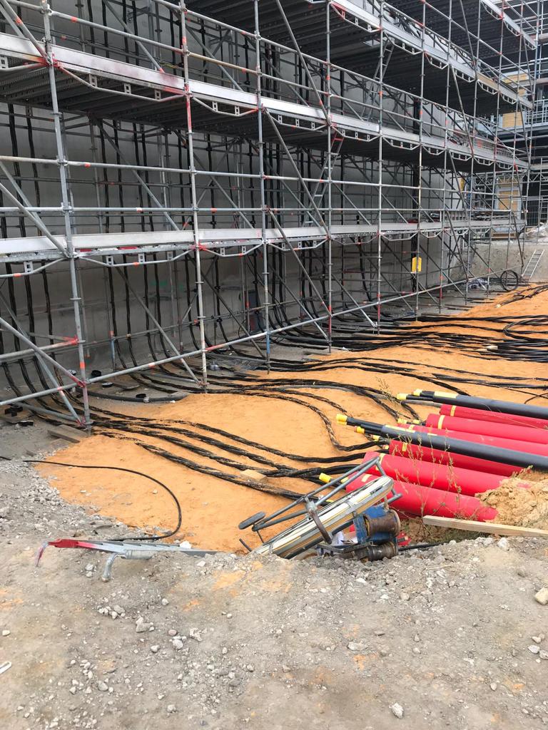 Großes Baugerüst an einer Gebäudewand, davor zahlreiche schwarze Kabelstränge und rote Schutzrohre, die auf sandigem Untergrund verlaufen, im Vordergrund liegen Bauwerkzeuge und Erdmaterial.