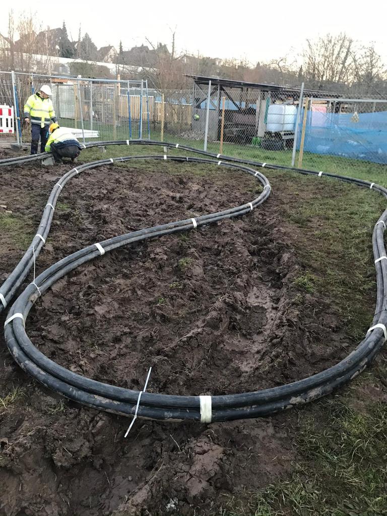 Zwei Arbeiter in Warnwesten und Helmen installieren auf einer matschigen Baustelle dicke schwarze Rohre, die in einer geschwungenen S-Form auf dem Boden ausgelegt und mit weißen Bändern fixiert sind.