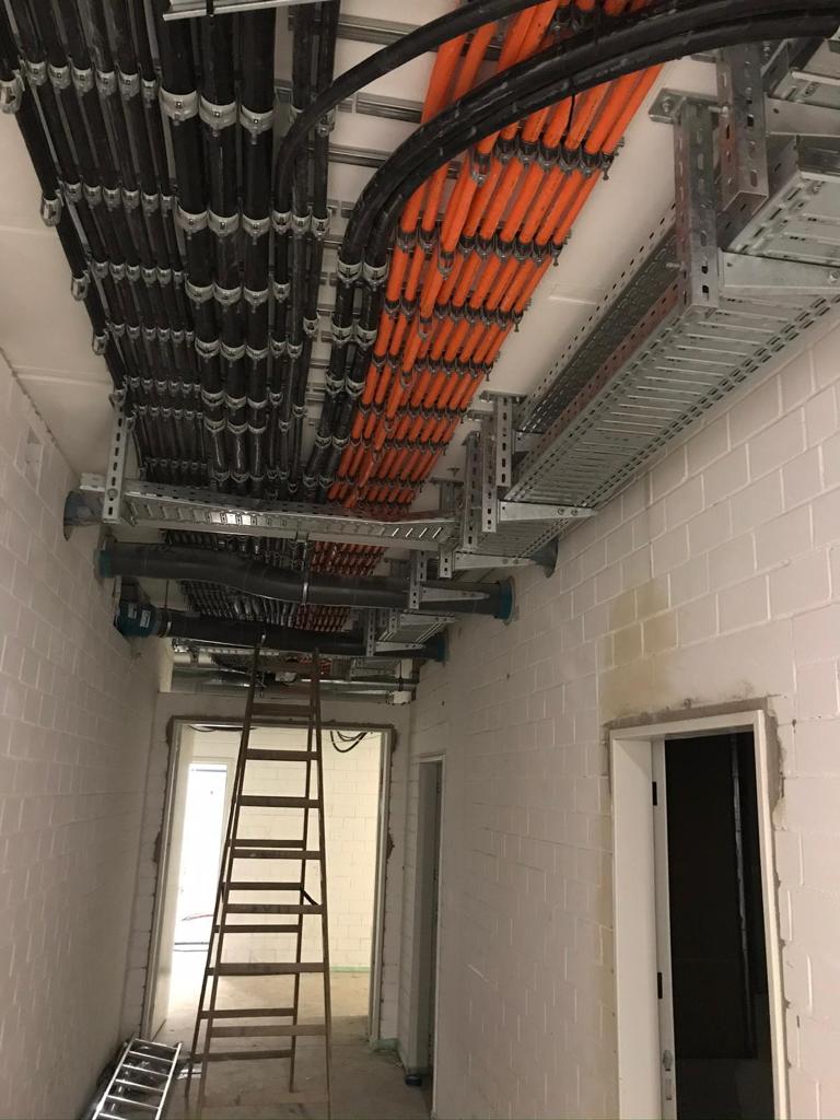 Blick in einen schmalen Flur mit weißen Wänden, an dessen Decke zahlreiche schwarze und orangefarbene Kabel in Kabeltrassen verlaufen. Eine Holzleiter steht mittig im Gang.